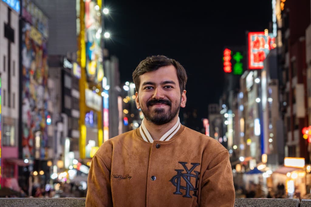 Pranav Jain in a suede varsity jacket, neon signage blurred behind him at night